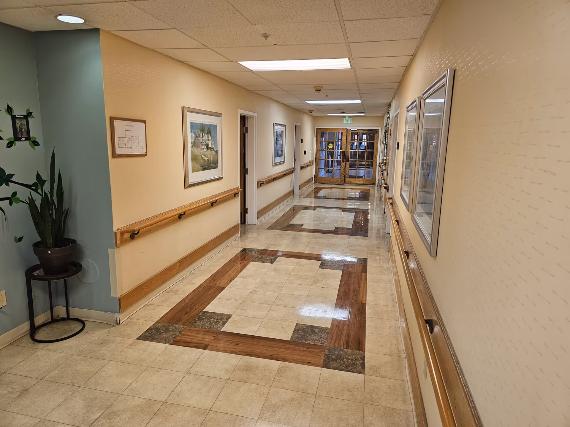 Well-lit interior hallway with handrails, framed artwork, patterned tile floor and double doors at the end.