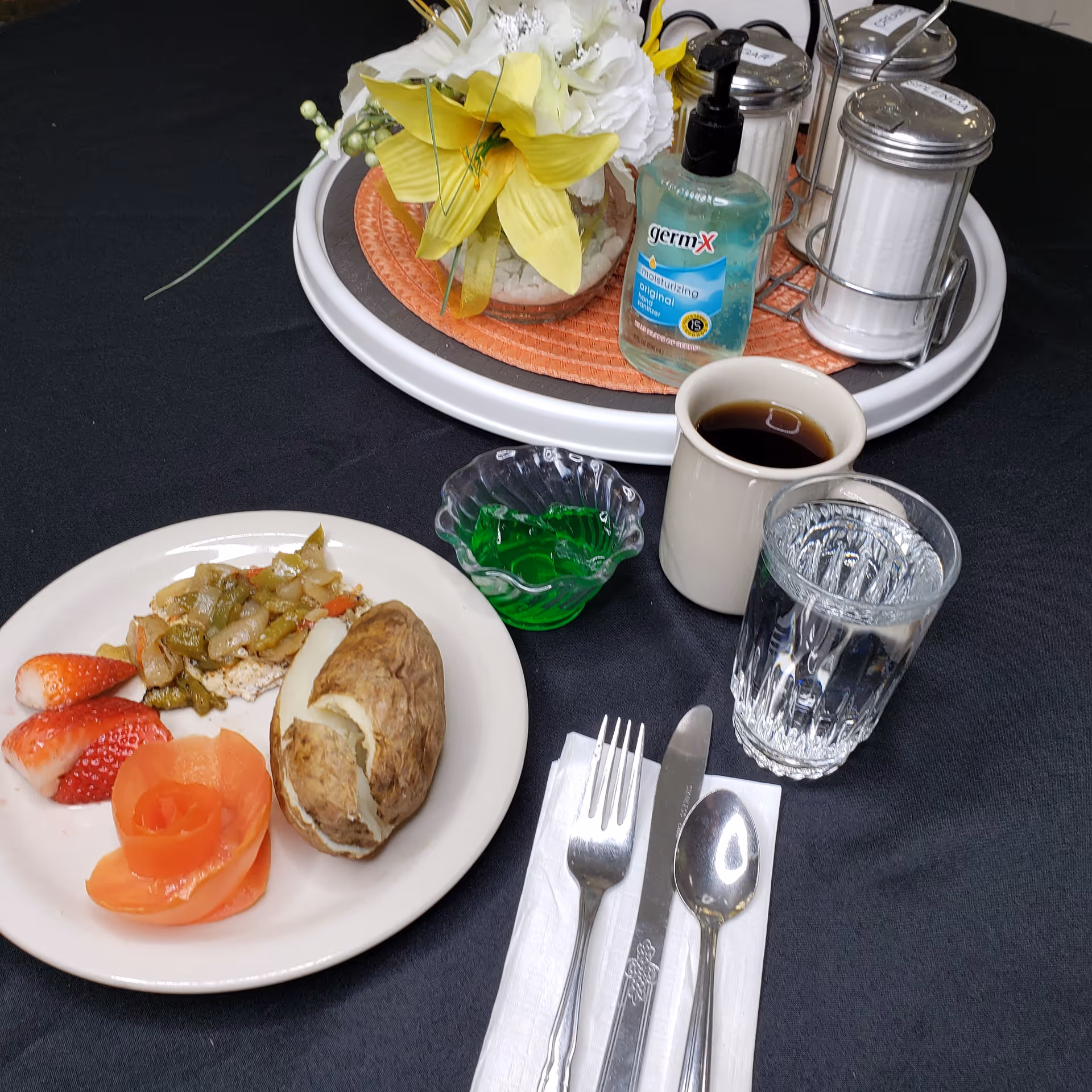 A meal served on a white plate with a baked potato, cooked vegetables, sliced strawberries, and a tomato rose. Next to the plate are a fork, knife, and spoon on a napkin, a glass of water, a cup of black coffee, and a small dish of green gelatin. In the background, there is a round tray with a floral arrangement, a bottle of Germ-X hand sanitizer, and condiment containers on a black tablecloth.