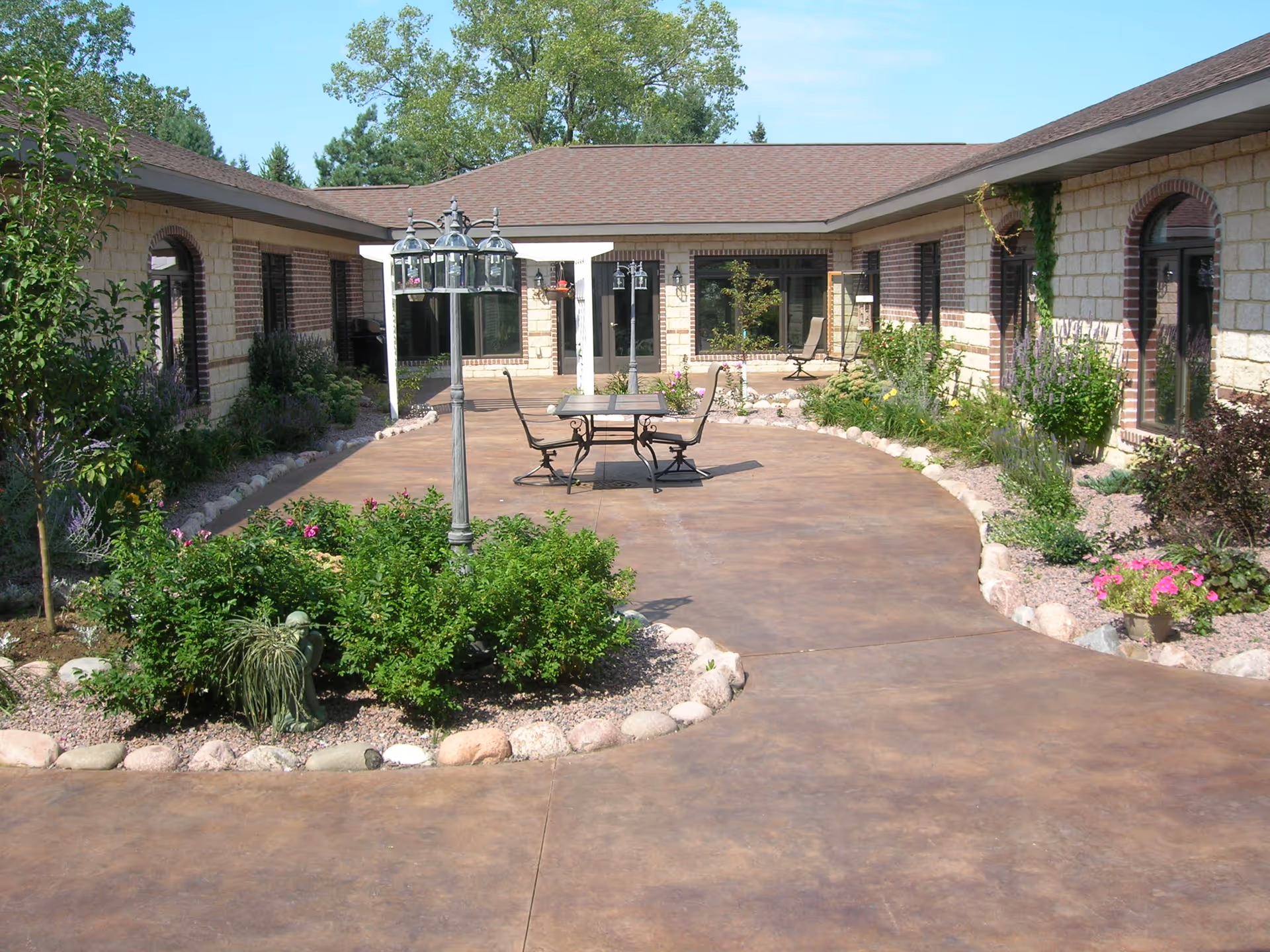 Sunlit courtyard with a paved walkway, central planted island, lamp post and a table with chairs surrounded by a single-story brick building.