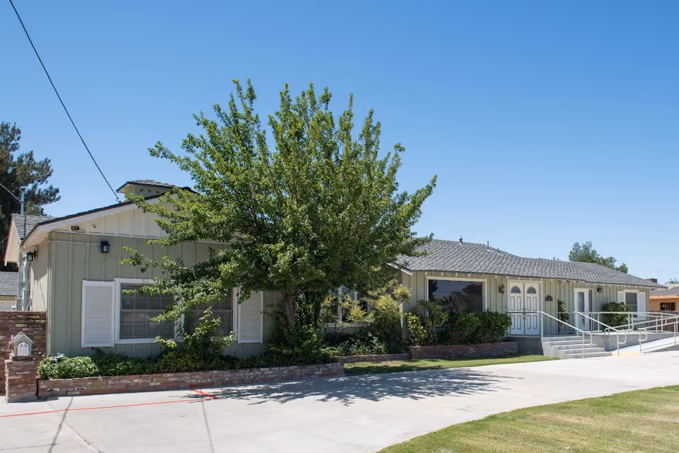 Exterior view of a single-story building with light green siding and white trim, featuring a large tree and shrubs in front. There is a concrete driveway and a ramp with railings leading to double doors under a clear blue sky.