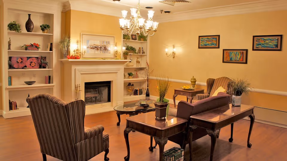Cozy assisted-living common room with a fireplace, built-in bookshelves, chandelier, armchairs, and wooden tables.
