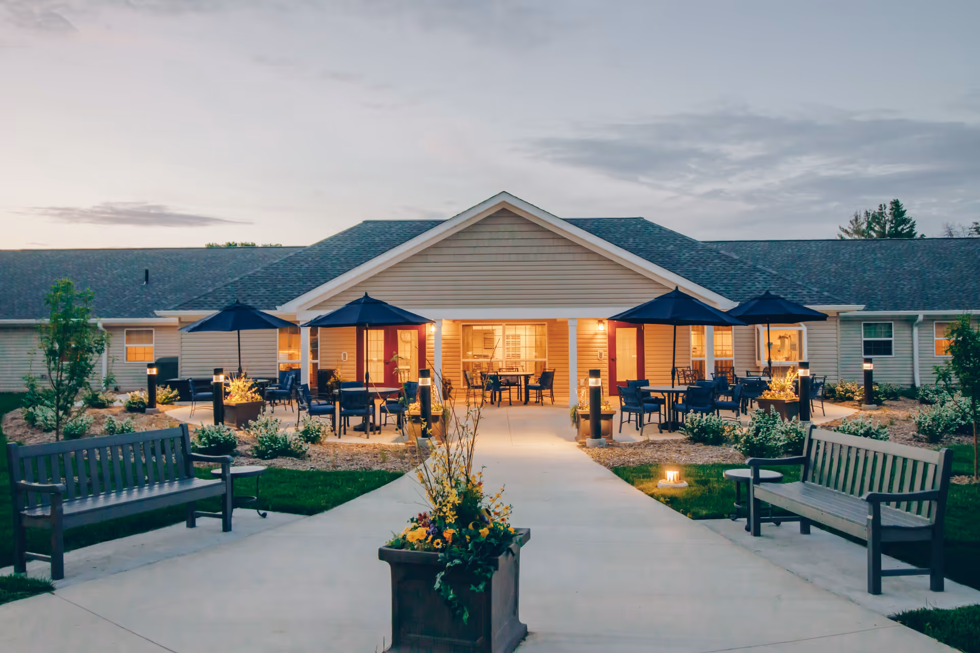 Outdoor patio area of a senior living facility at dusk with benches, tables, chairs, and umbrellas arranged on a paved walkway leading to the building entrance, surrounded by landscaped plants and illuminated by soft lighting.