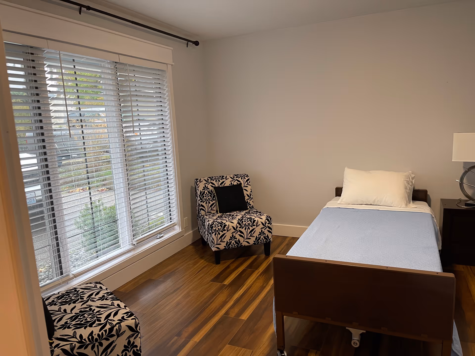 A simple bedroom with a single bed made with a white pillow and light blue blanket. There are two patterned chairs with black and white floral designs, one near a large window with white blinds and the other partially visible. The floor is wooden, and there is a small dark nightstand with a lamp on it next to the bed.