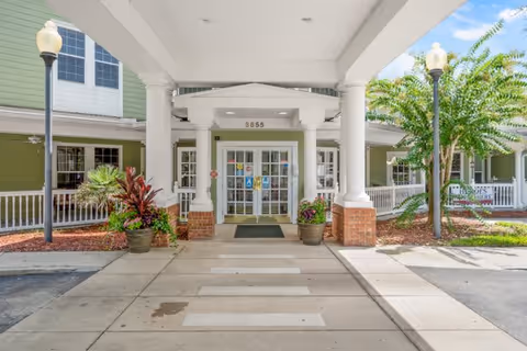 Covered porte-cochère and main entrance with glass doors, columns, potted plants, and a paved walkway.