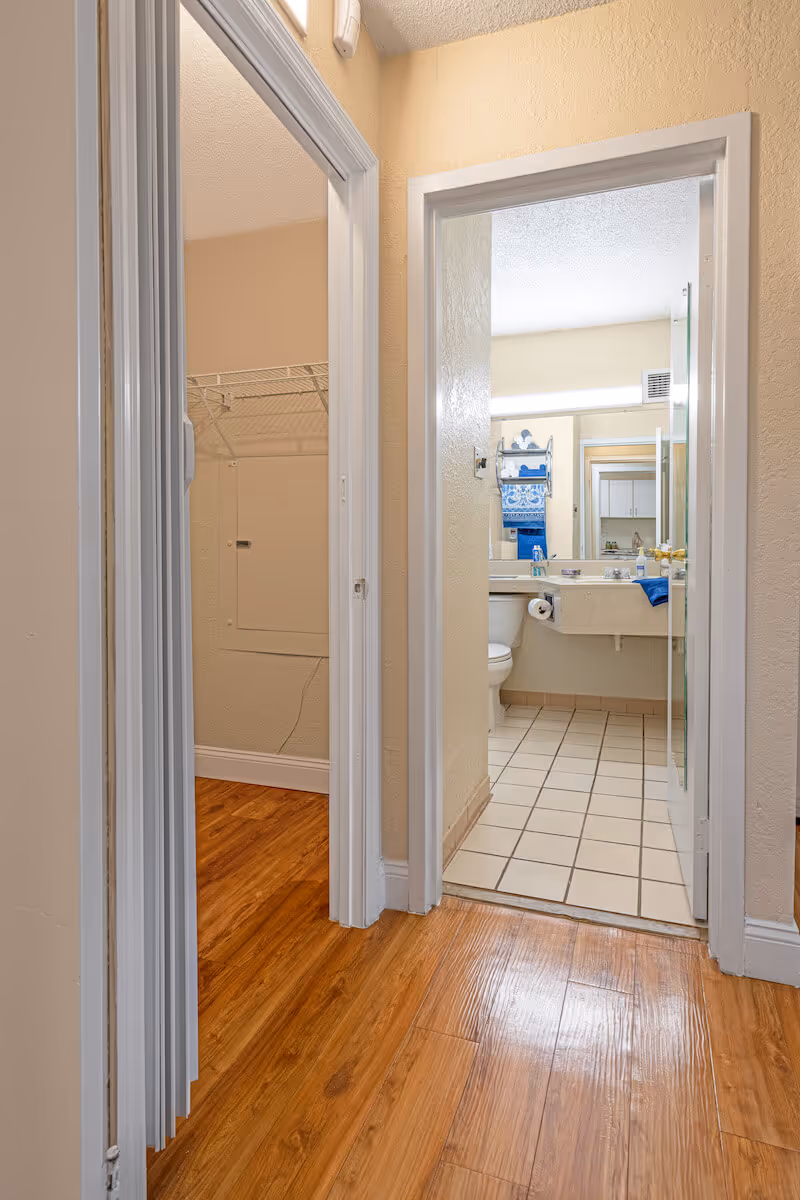 View down a hallway with wood flooring leading to a bathroom with tiled floor. The bathroom has a sink with a large mirror, a toilet, and blue towels hanging on a rack. To the left of the hallway is an open closet with a wire shelf.