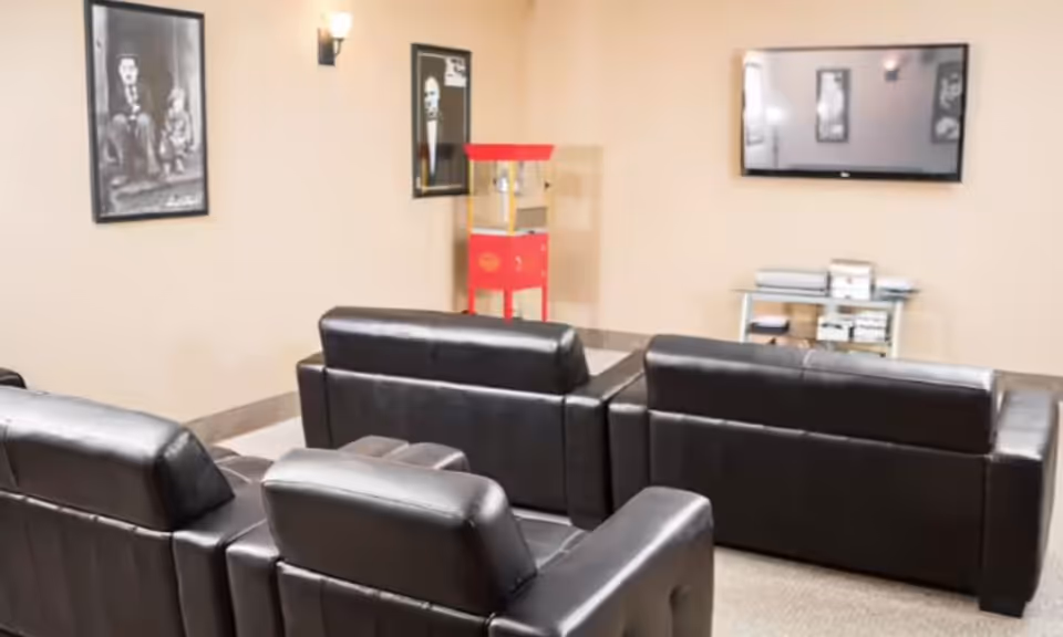 A cozy entertainment room with four black leather sofas arranged in two rows facing a wall-mounted flat-screen TV. On the wall are two framed black and white portraits, and a red popcorn machine stands in the corner. Below the TV is a small metal shelf holding electronic devices.