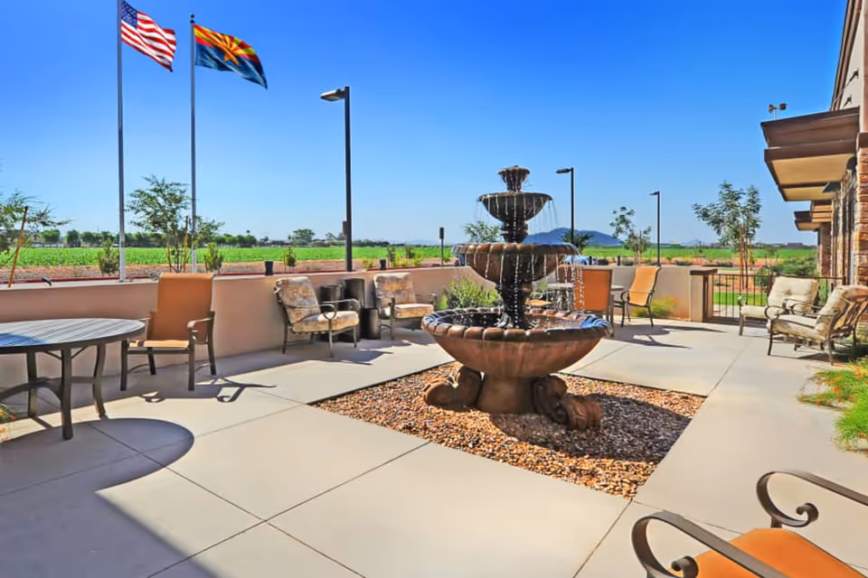 Outdoor patio area at Sky Ridge facility with a three-tiered water fountain in the center, surrounded by chairs and tables. Three flagpoles with the American flag, Arizona state flag, and another flag are visible against a clear blue sky. The patio overlooks a green field with distant mountains.