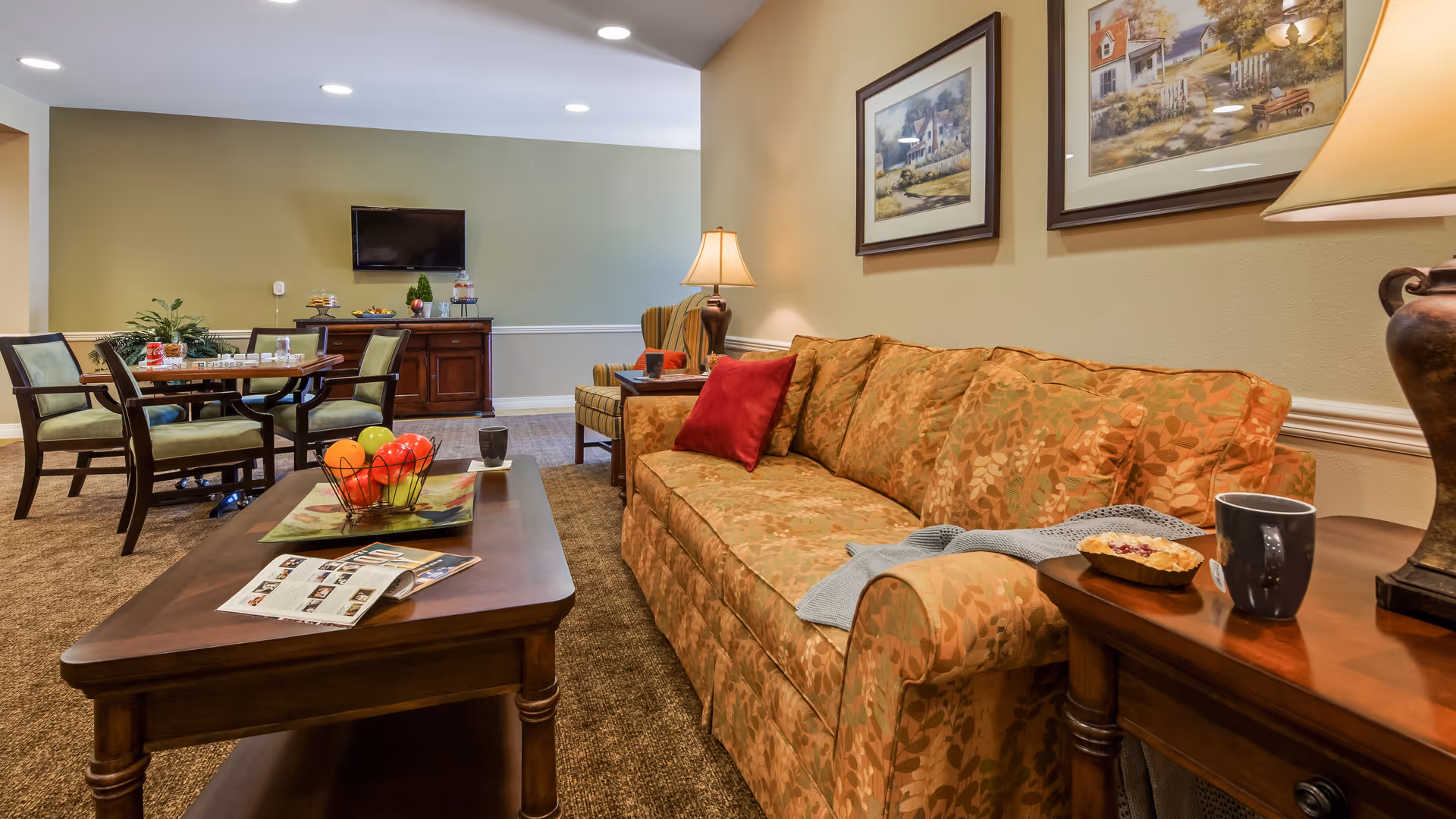A cozy living room area in an assisted living facility featuring a floral patterned sofa with a red pillow and a gray throw, a wooden coffee table with a fruit basket and magazines, a side table with a lamp, a cup, and a small pie. In the background, there is a dining table with chairs, a TV mounted on a green wall, and framed paintings hanging above the sofa.