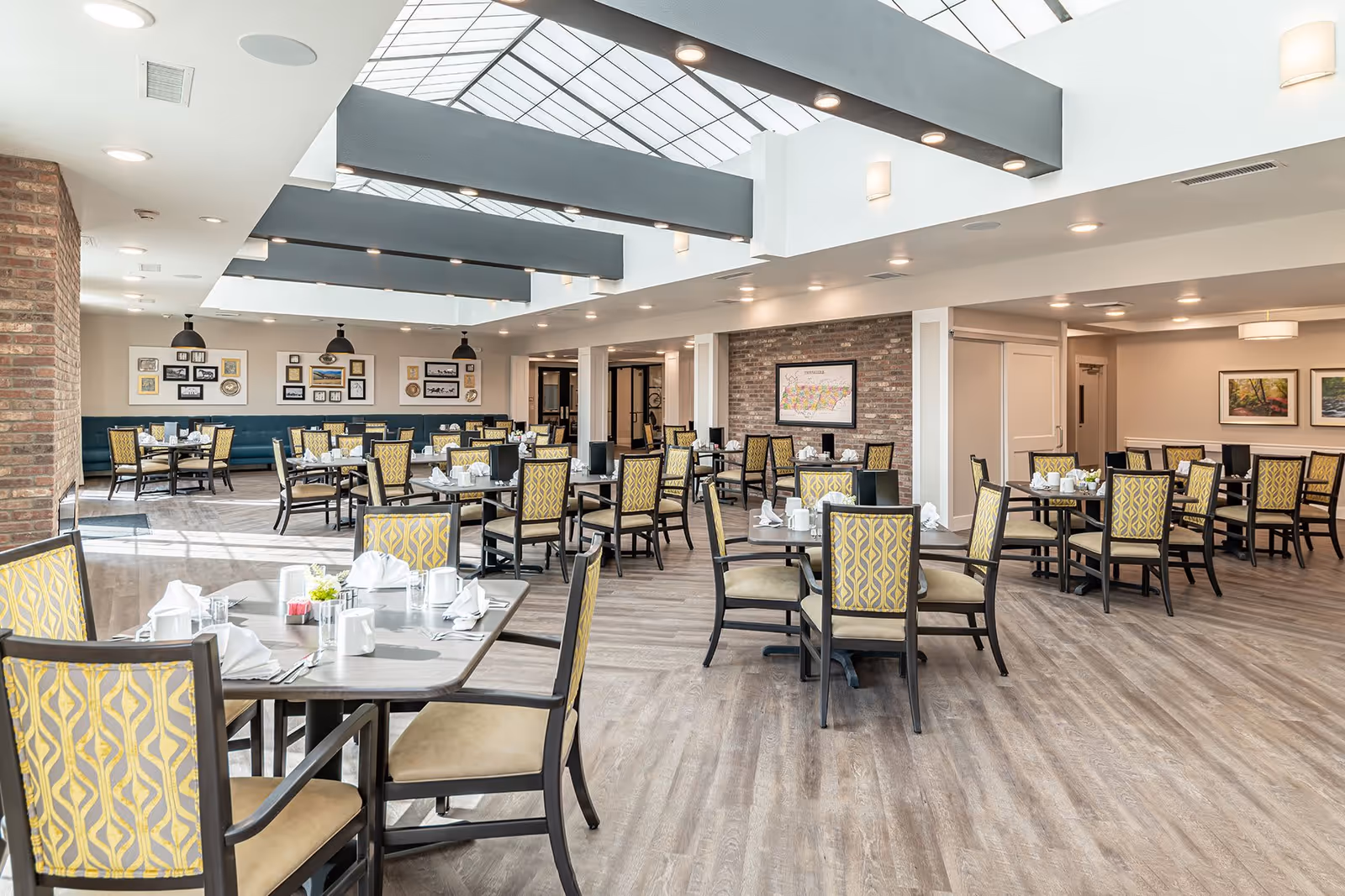 A spacious and well-lit dining room with multiple tables and chairs arranged neatly. The chairs have yellow patterned upholstery, and the tables are set with white napkins, cups, and silverware. The room features a large skylight with exposed beams, brick accent walls, framed artwork, and wood flooring.