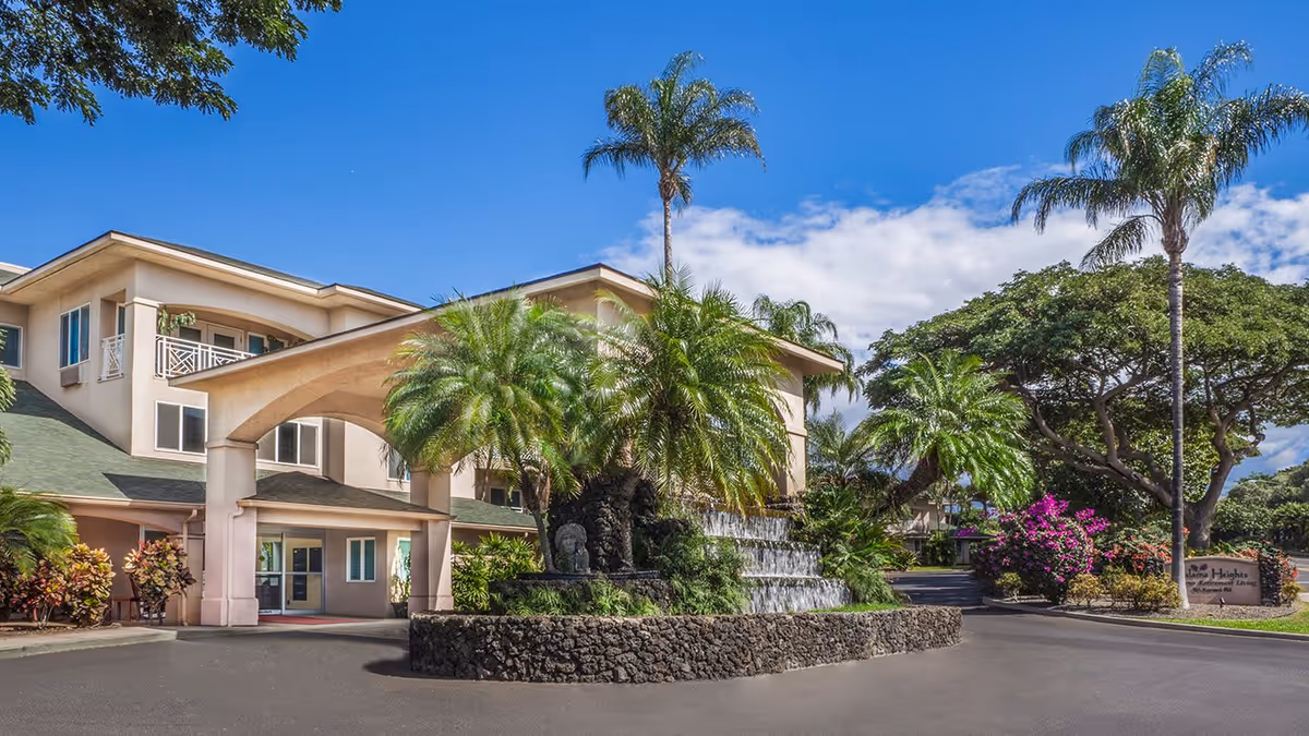 Exterior view of Holiday Kalama Heights facility featuring a beige multi-story building with a covered entrance, surrounded by palm trees, lush greenery, and a tiered stone waterfall feature under a clear blue sky.