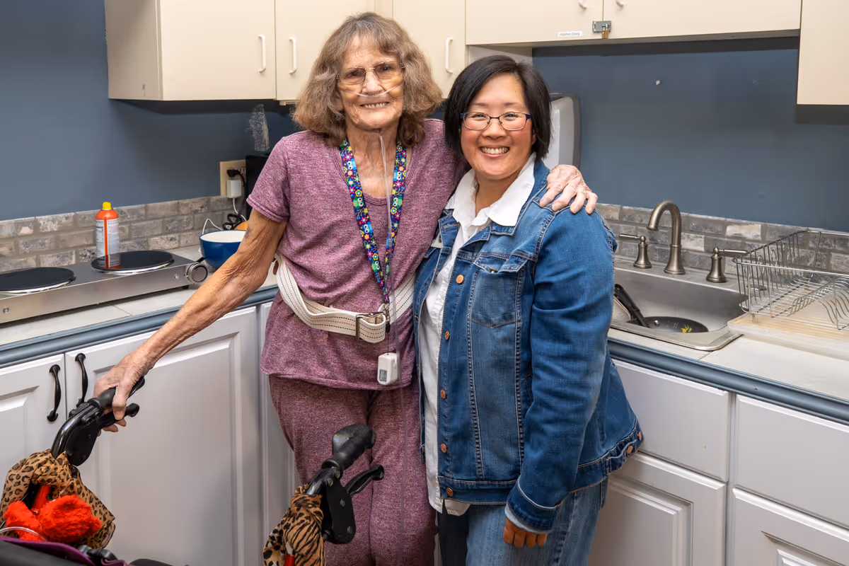 An elderly woman using a walker and a younger woman standing together in a kitchen, both smiling. The kitchen has white cabinets, a sink, and a countertop with a hot plate and some kitchen items.