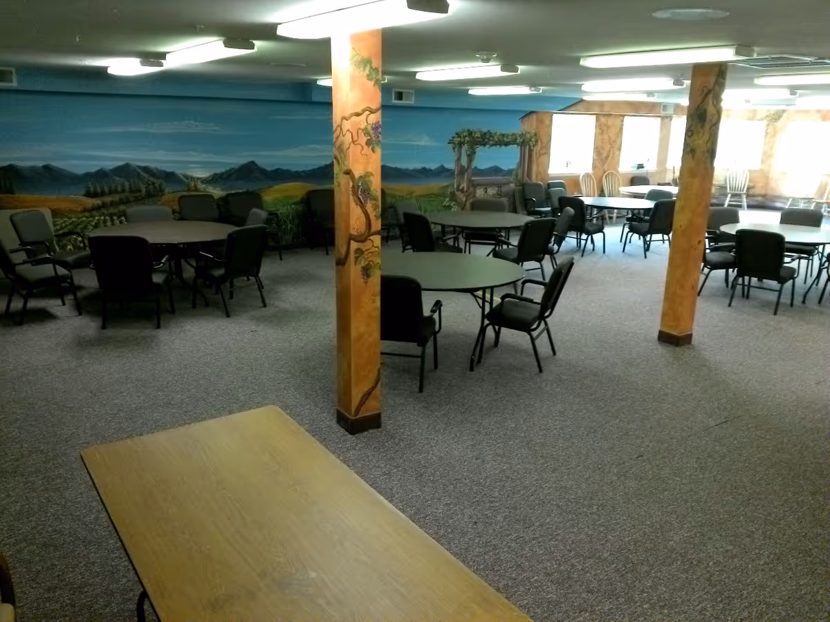 A large room with multiple round tables and chairs arranged for group seating. The walls are decorated with a mural depicting a scenic landscape with mountains, fields, and a pergola. The room has carpeted flooring and several painted support columns.
