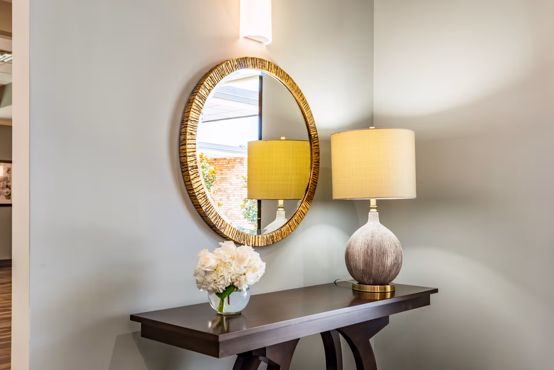 Console table with a round mirror, a table lamp, and a small vase of flowers against a pale wall.