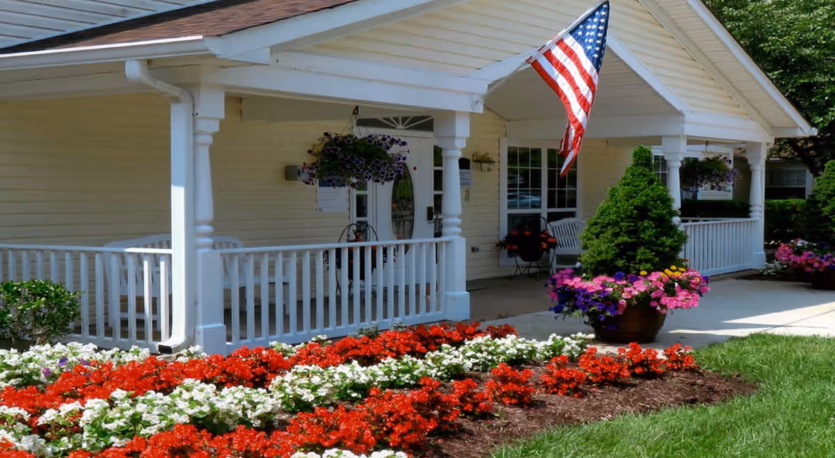 Front porch of a yellow building with white railings and columns, decorated with hanging flower baskets and potted plants. An American flag is displayed near the entrance, and there are red and white flowers planted in the garden bed in front of the porch.