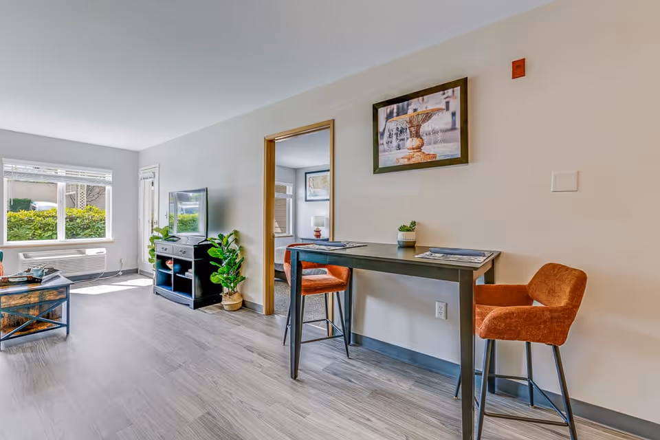 A bright and modern living area in a senior living facility featuring a window with a view of greenery, a TV on a black stand, a small table with two orange chairs, a framed picture of a fountain on the wall, and a doorway leading to a bedroom.