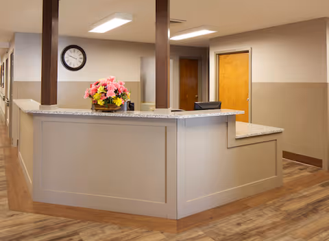 Reception desk area with a flower arrangement, wall clock, and hallway doors in a senior living facility.