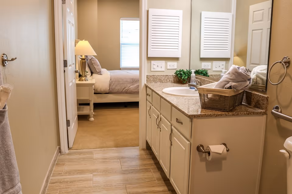 View of a bathroom with a granite countertop, sink, and a basket with rolled towels. The bathroom has beige walls, a towel ring, and a toilet paper holder. Through an open door, a bedroom with a bed, nightstand, lamp, and window with blinds is visible.