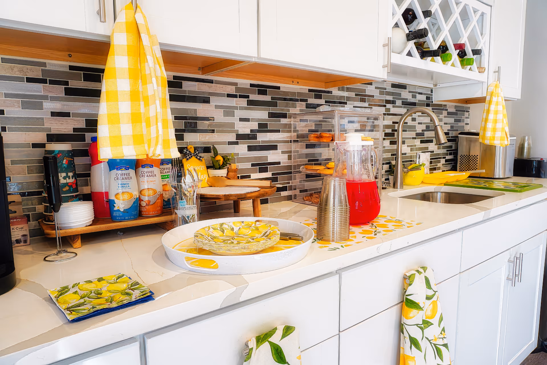 A bright kitchen countertop with white cabinets and a modern faucet. The backsplash features a mosaic of gray, black, and beige tiles. On the counter, there are yellow and white checkered towels hanging, coffee creamers, a glass pitcher with red liquid, stacked plastic cups, a tiered tray with pastries, and lemon-themed plates and napkins. A wine rack with several bottles is mounted above the counter.