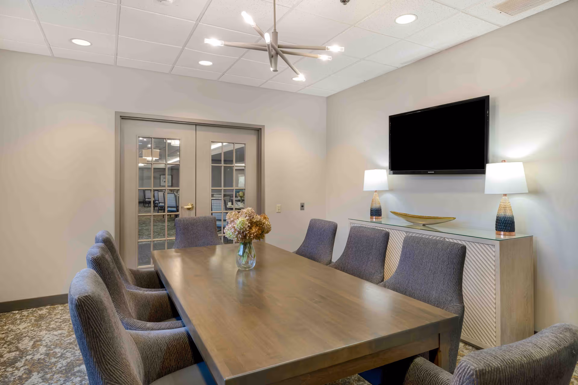 A modern meeting or dining room with a long wooden table surrounded by eight upholstered chairs. On the table is a glass vase with flowers. The room has beige walls, a patterned carpet, and a ceiling light fixture with multiple bulbs. At the far end, there are double glass doors and a sideboard with two table lamps and a decorative bowl, with a flat-screen TV mounted on the wall above it.