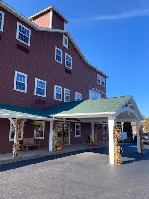 Exterior view of a multi-story building with red siding and white trim, featuring a covered entrance with a green roof. The entrance is decorated with hanging flower pots and autumn-themed decorations including pumpkins and dried corn stalks. The sky is clear and blue.