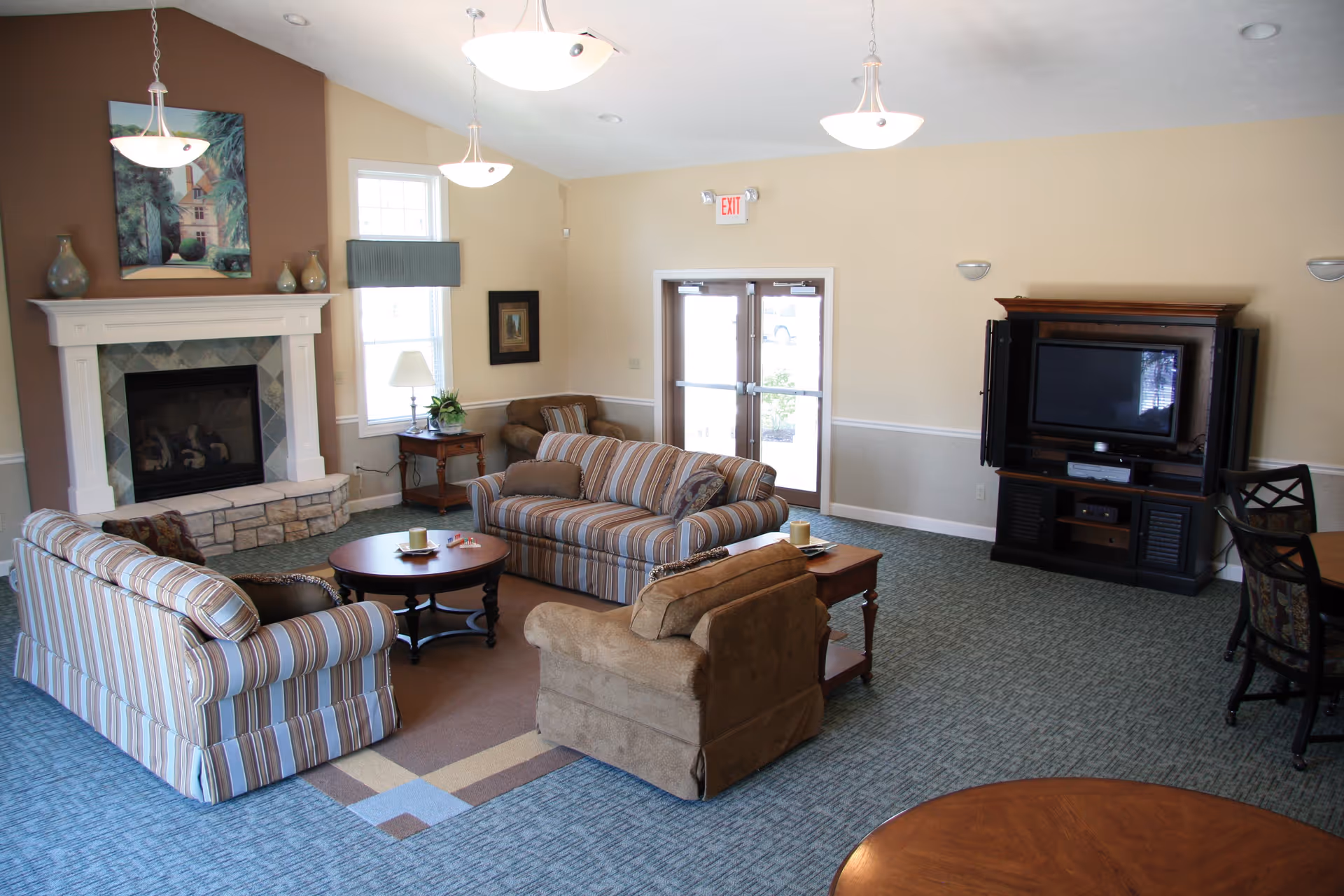 A cozy living room area with two striped sofas, one brown armchair, a round wooden coffee table, and a fireplace with a painting above it. There is a wooden entertainment center with a TV and a glass door leading outside. The room has beige walls, carpeted floors, and hanging ceiling lights.