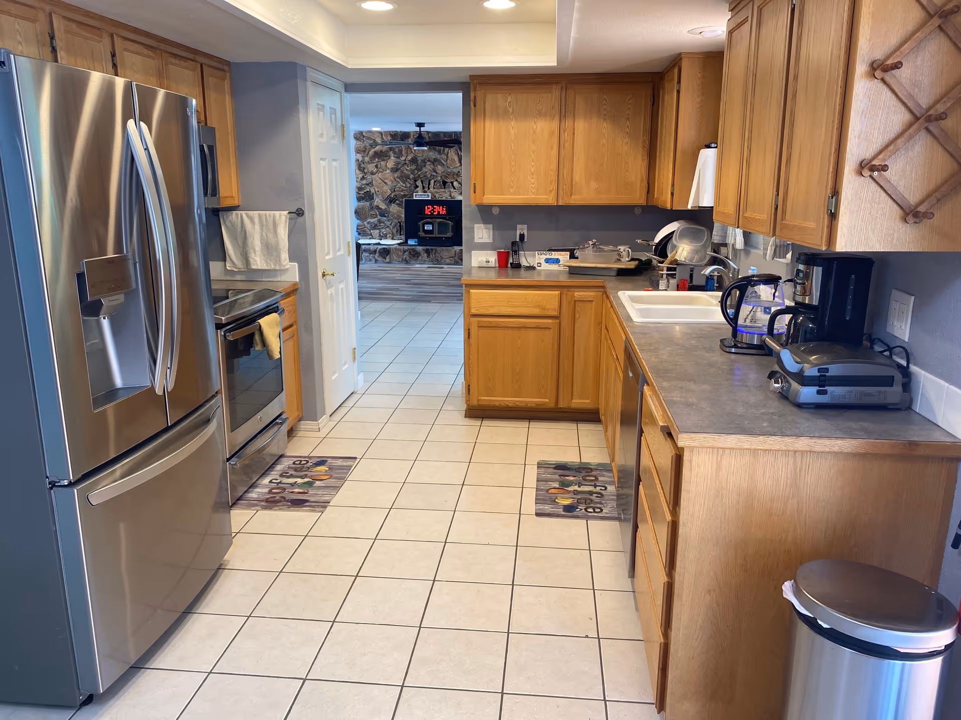 A kitchen with wooden cabinets, a stainless steel refrigerator, an oven, a sink, a coffee maker, a kettle, and a sandwich press on the countertop. The floor is tiled, and there are two small rugs with coffee cup designs. In the background, there is a doorway leading to a room with a stone fireplace and a digital clock showing 12:34.