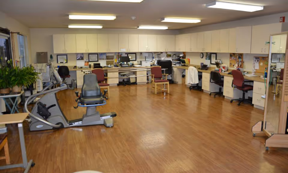 A spacious room with wooden flooring featuring exercise equipment in the foreground and a long counter with multiple office chairs, computers, and cabinets along the back wall. The room is well-lit with ceiling lights and has some plants on the left side near the window.