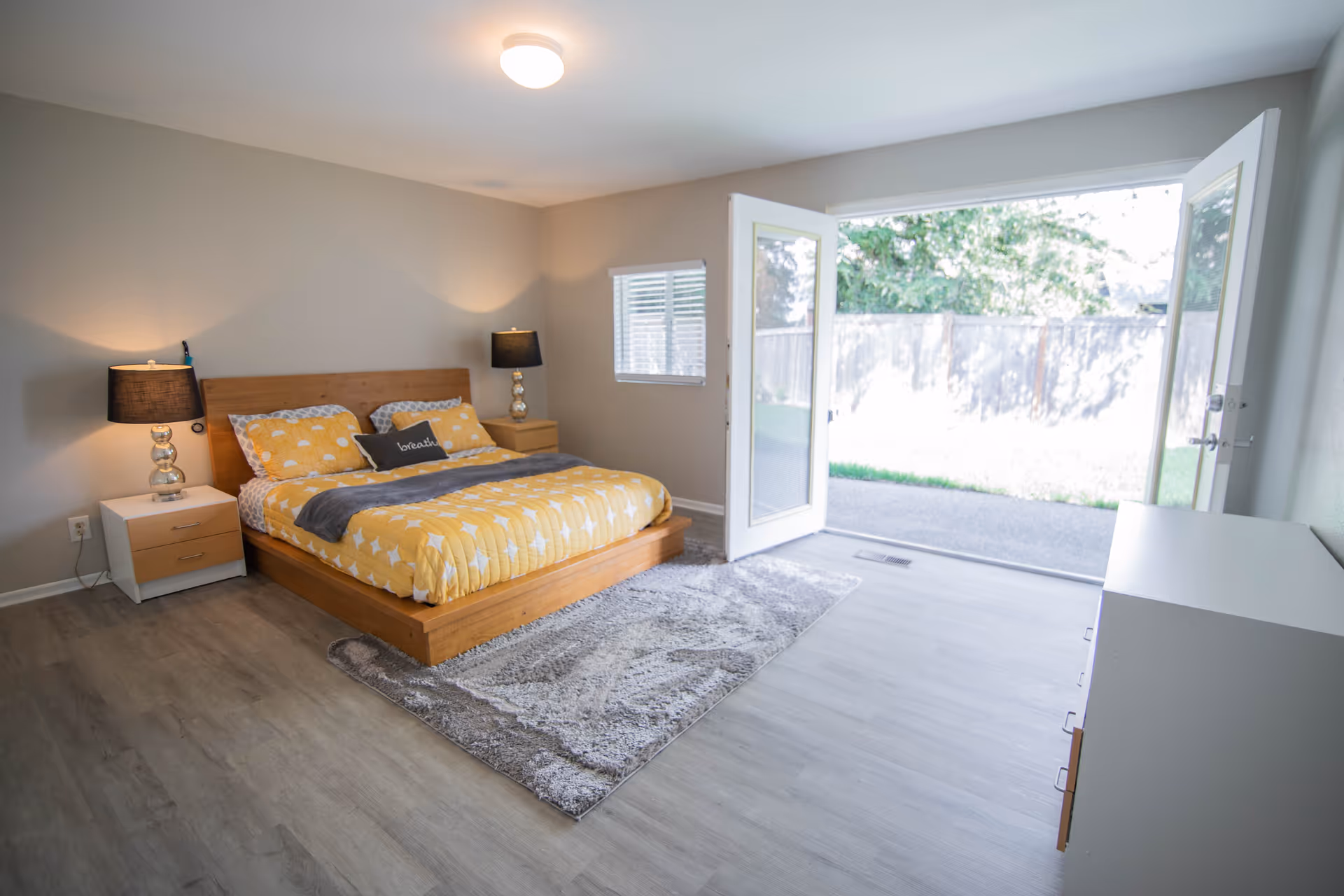 Bright bedroom with a wooden platform bed dressed in yellow bedding, nightstands with lamps, and open double doors leading to a backyard.
