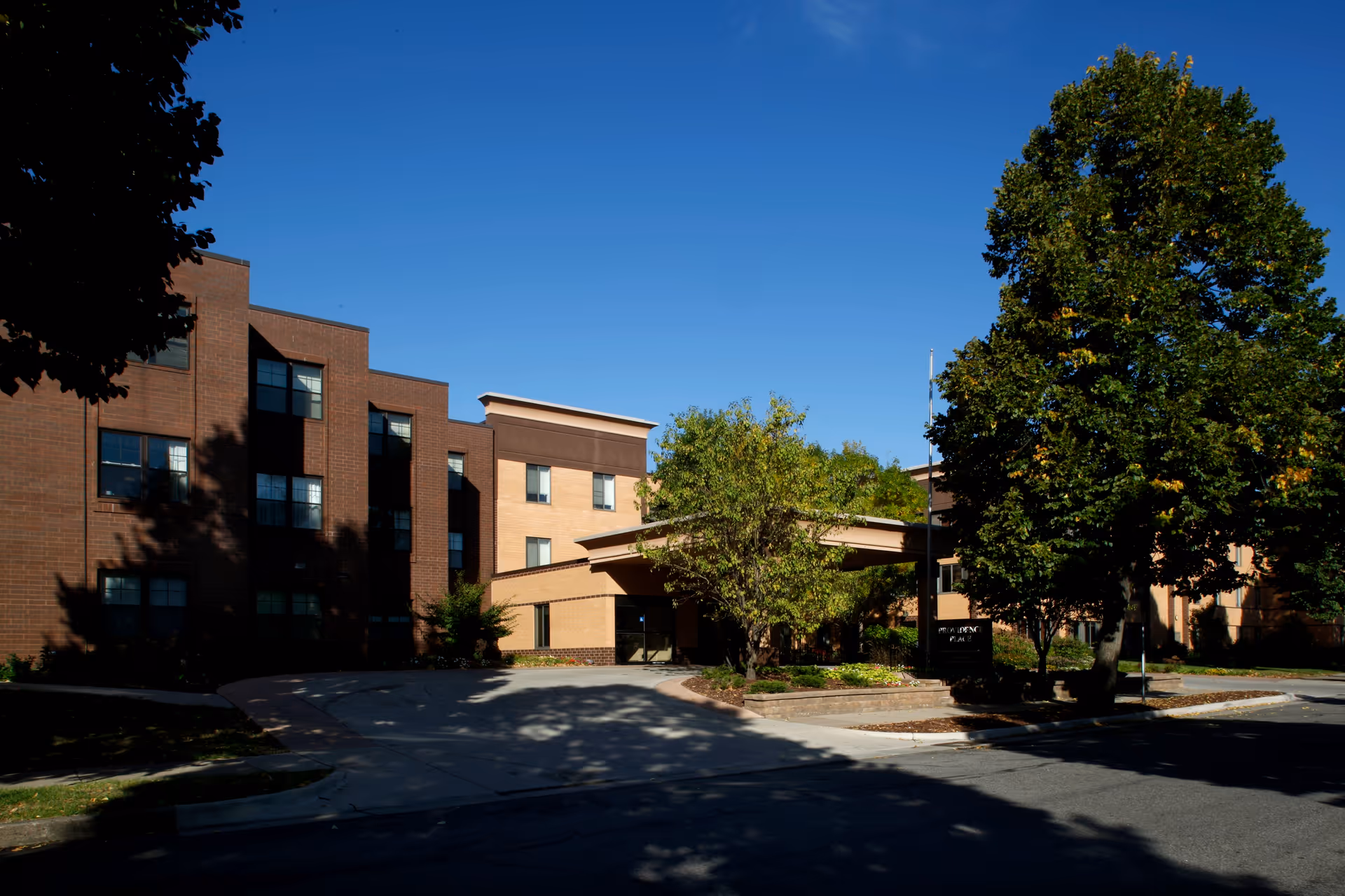 Front exterior of a multi-story brick senior living building with trees, a covered entrance and driveway under a clear blue sky.