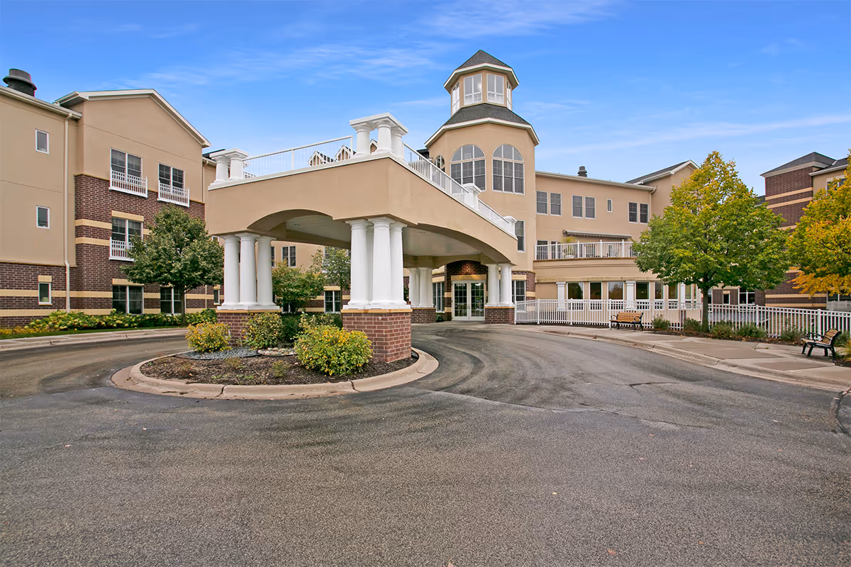 Exterior view of Boutwells Landing senior living facility showing a large covered entrance with white columns, a circular driveway, landscaped bushes, trees, and a multi-story building with beige and brick facade under a clear blue sky.