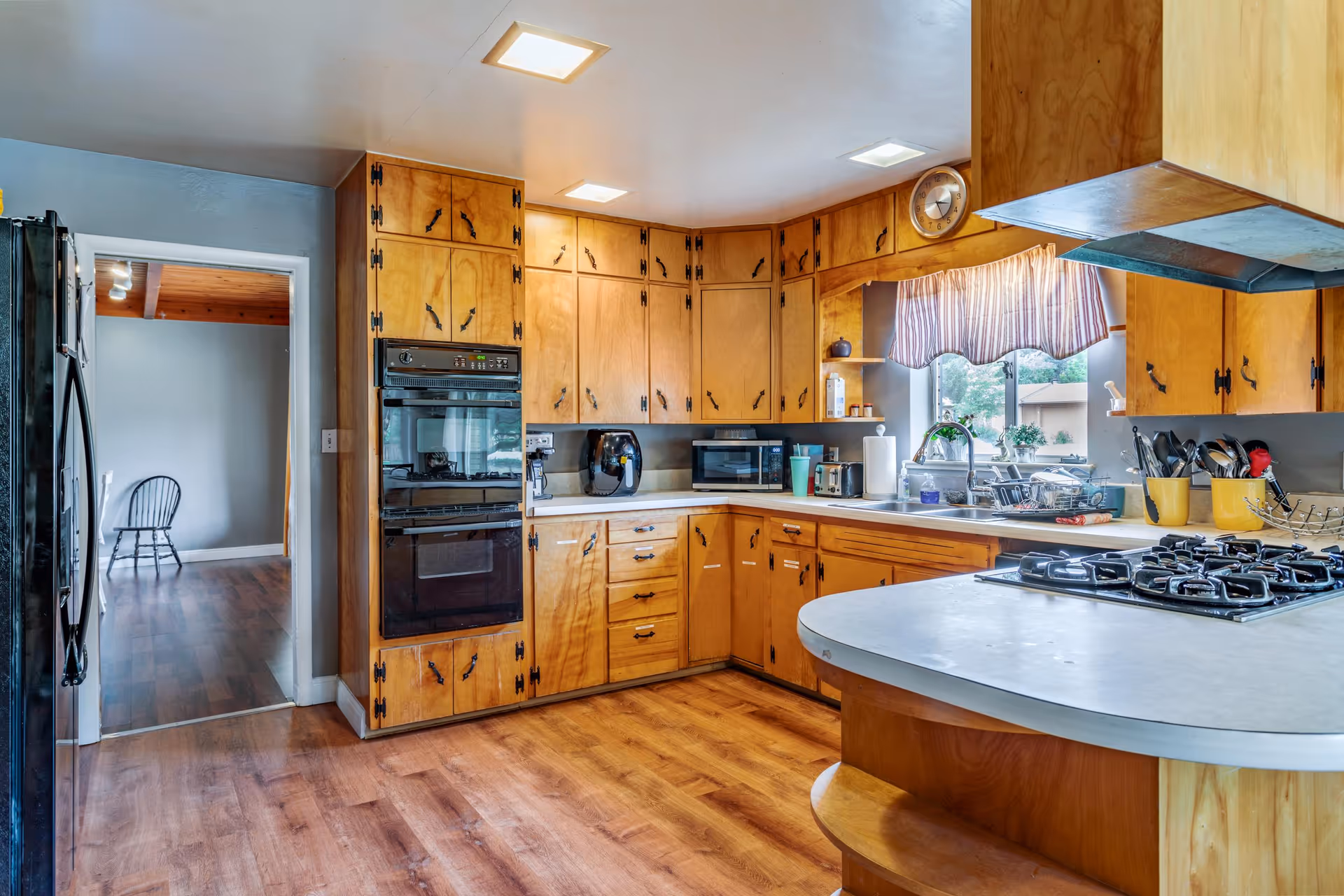 A kitchen with wooden cabinets and drawers, a gas stove on a curved countertop, a double oven, a microwave, an air fryer, and a coffee maker. There is a window with a striped valance above the sink, and various kitchen utensils and appliances are placed on the counters. The floor is wooden, and there is a doorway leading to another room with a wooden chair visible.