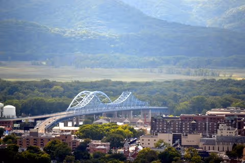 A scenic view of a cityscape with a large arched bridge spanning across a river or valley, surrounded by buildings and lush green trees, with forested hills and mountains in the background under a clear sky.