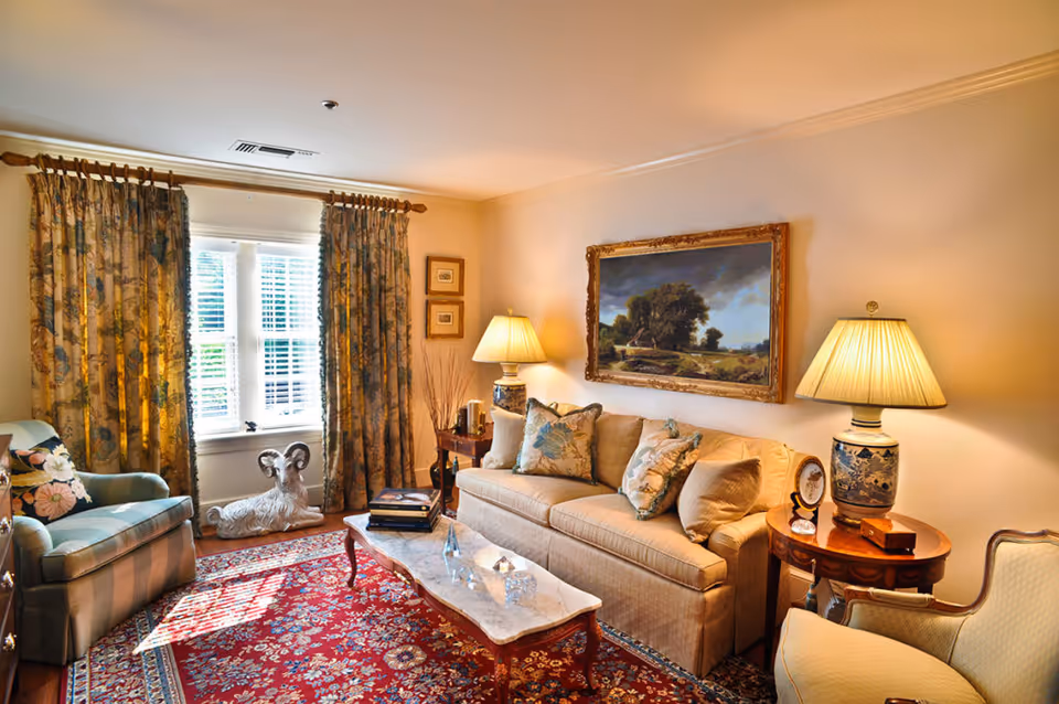 A cozy living room with a beige sofa adorned with floral pillows, a marble coffee table with books and decorative items, two side tables each with a lamp, an armchair with striped upholstery, floral curtains framing a window with white shutters, a red patterned rug, and a large framed landscape painting on the wall.