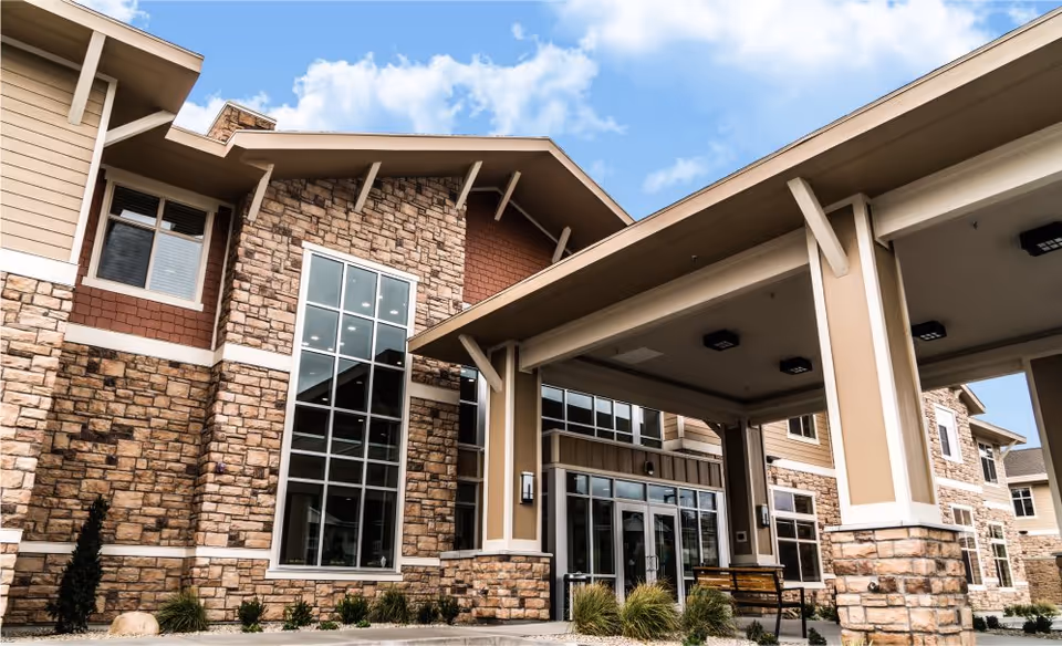 Exterior view of a senior living facility building with stone and beige siding, large windows, and a covered entrance area under a blue sky with some clouds.