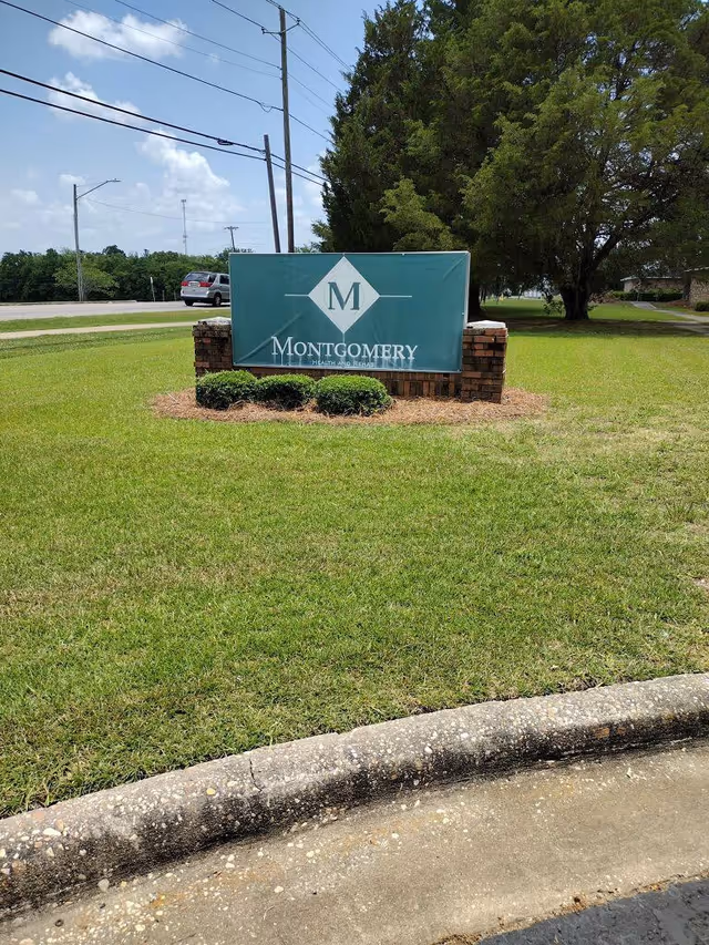 A green Montgomery Health and Rehab entrance sign on a brick base in a grassy lawn by the road.