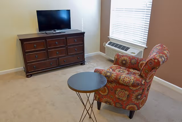 A cozy corner of a living room featuring a patterned armchair with red, yellow, and orange floral designs, a small round black table with thin metal legs, a wooden dresser with multiple drawers, and a flat-screen TV on top. There is a window with white blinds and an air conditioning unit below it. The walls are painted beige and light brown, and the floor is carpeted.