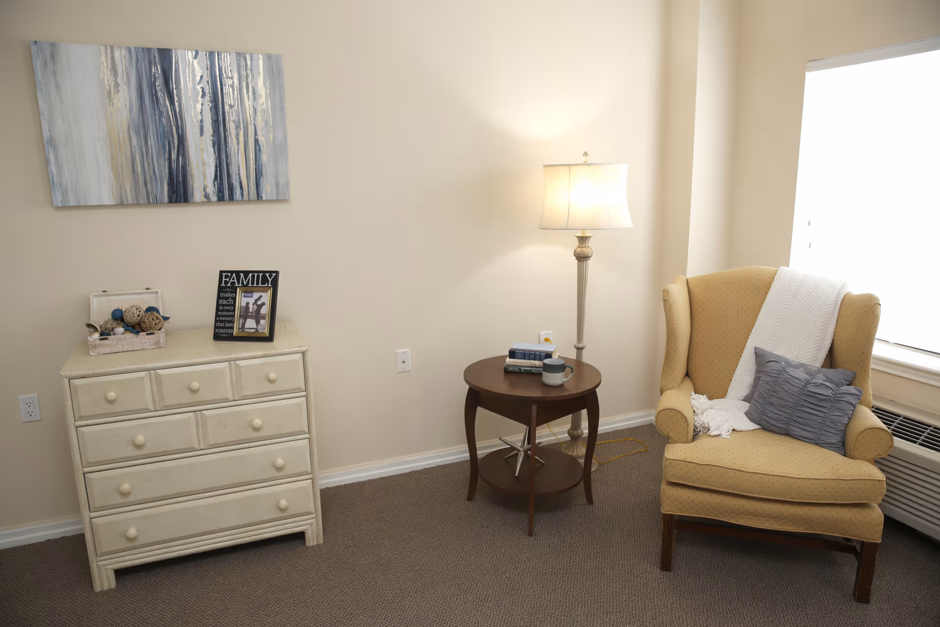 A cozy corner of a room featuring a beige wingback armchair with a gray pillow and white throw blanket, a round wooden side table with books and a mug, a tall floor lamp, and a cream-colored dresser with decorative items and a framed photo on top. A large window with blinds is partially visible on the right side.