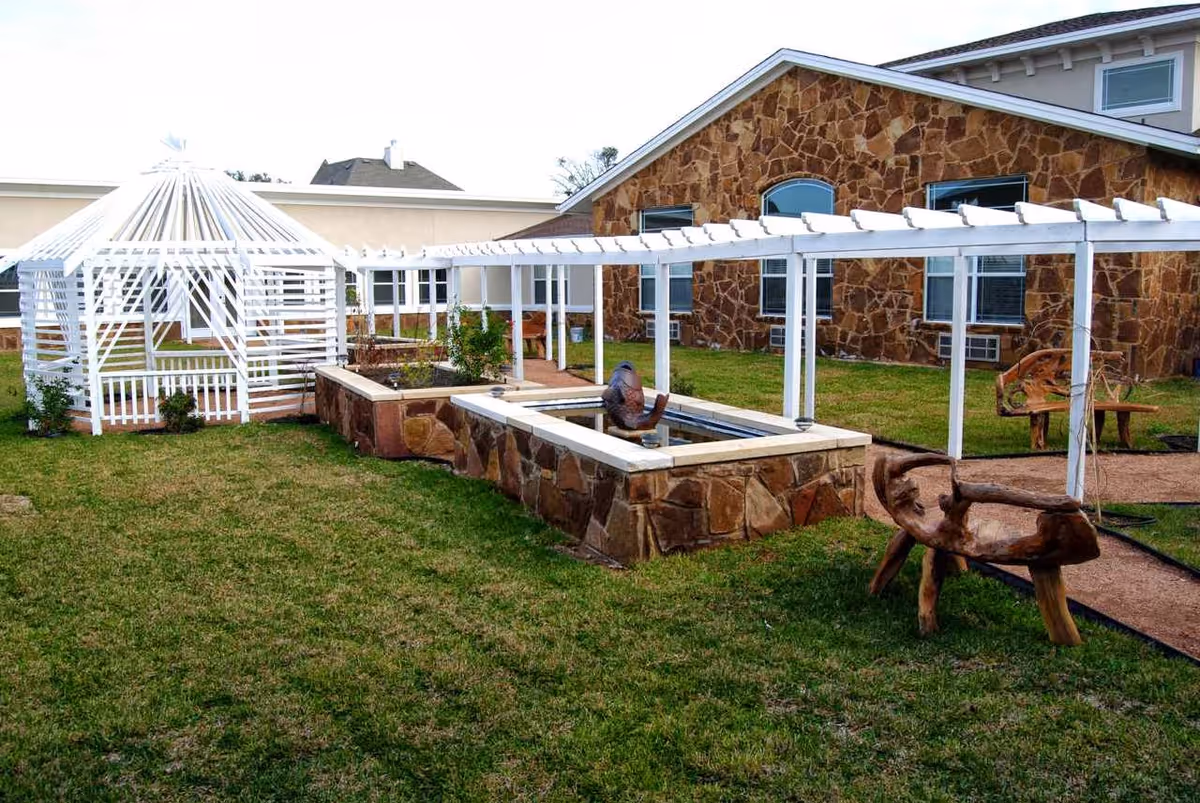 Outdoor garden area with green grass, a white wooden pergola structure, stone planters, a small water feature with a bird sculpture, and rustic wooden benches. The background shows a building with stone walls and windows.