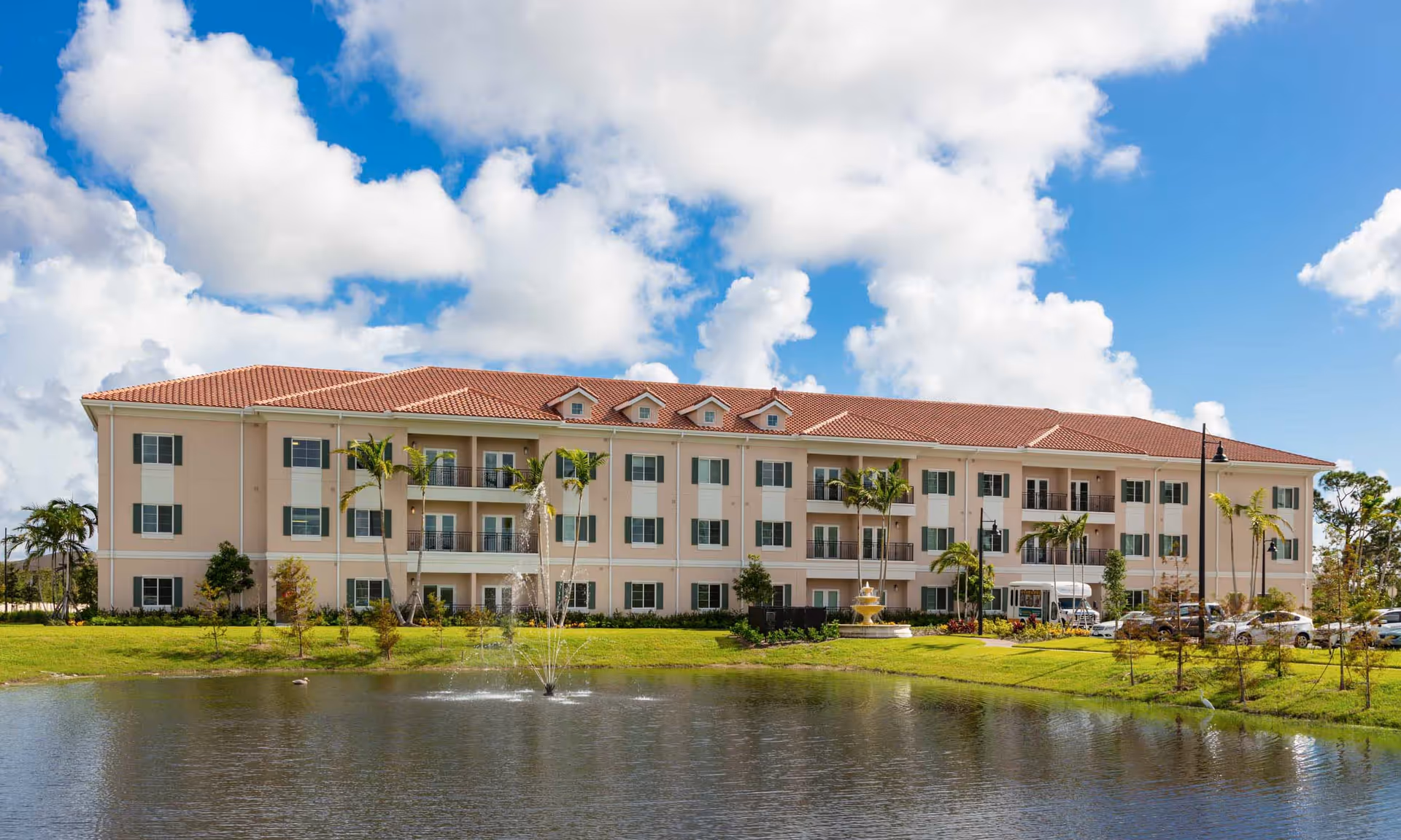 Three-story beige senior living building with a red tile roof and balconies facing a pond with a fountain under a blue sky.