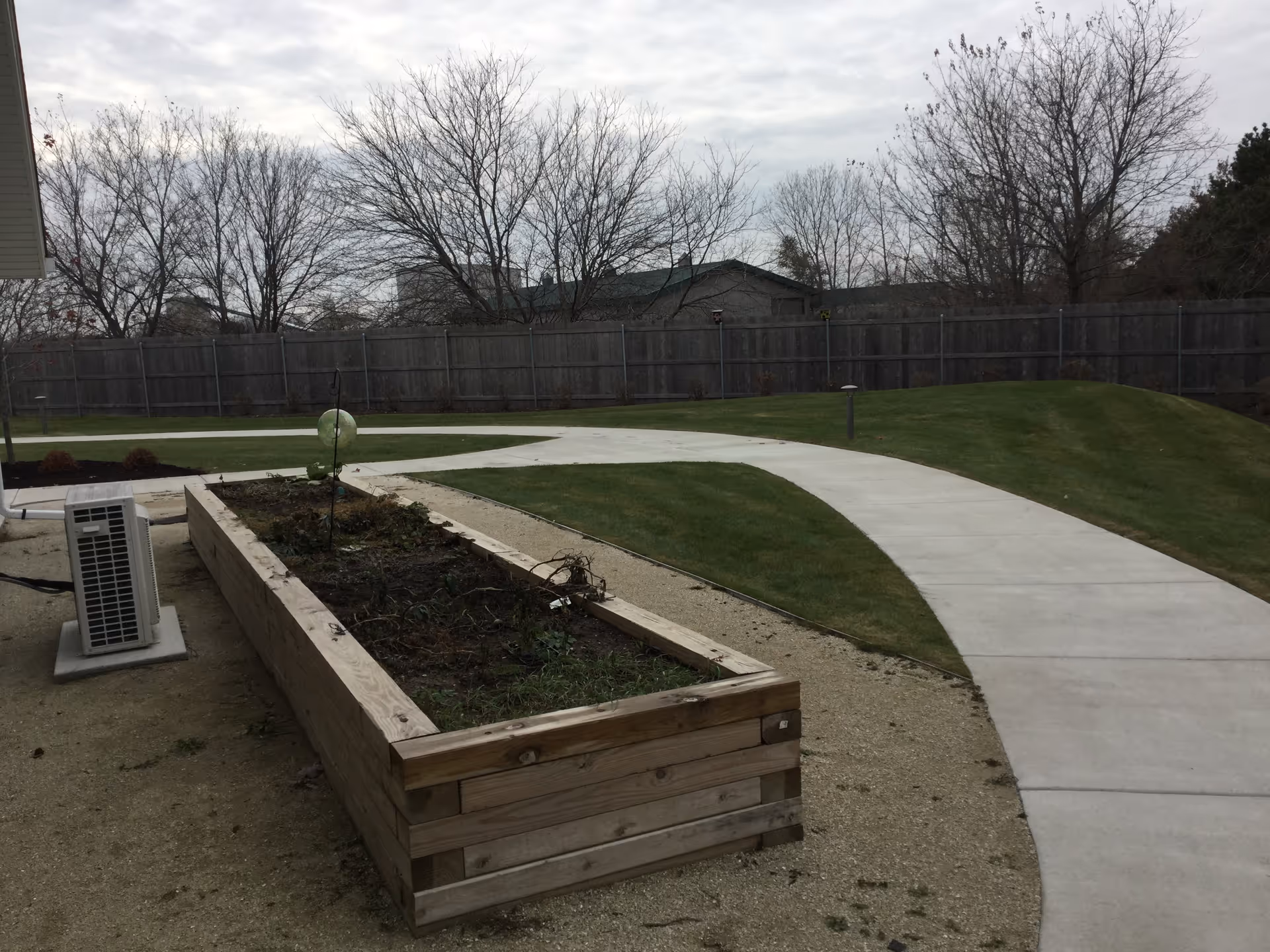 Outdoor garden area with a raised wooden planter box containing some plants, a curved concrete walkway, green grass, leafless trees, and a wooden fence in the background under a cloudy sky.