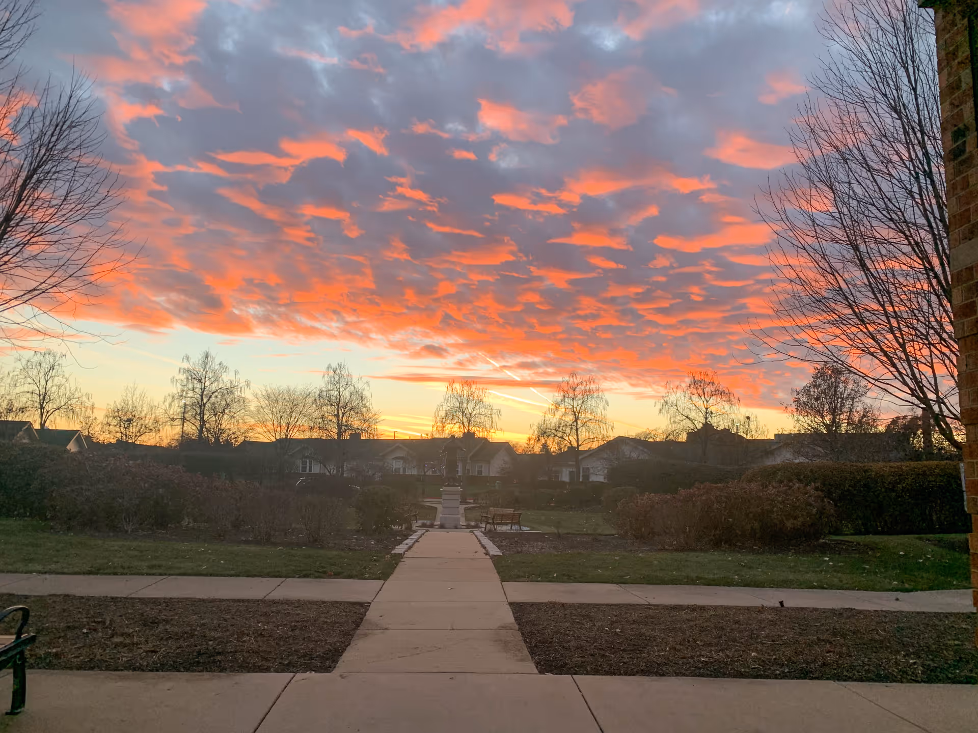 A scenic outdoor view at sunset with a paved walkway leading through a garden area with bushes and leafless trees. Residential buildings are visible in the background under a sky filled with dramatic orange and pink clouds.