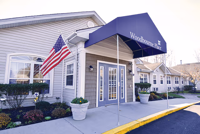 Exterior front entrance of a senior living facility with a blue awning labeled 'Woodbourne Place'. The building is light-colored with white trim, an American flag mounted on the wall, and landscaping with bushes and flowers near the entrance.