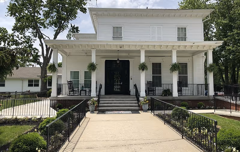 Front exterior view of a white two-story building with a covered porch featuring hanging plants and rocking chairs. There are steps and ramps leading up to the entrance with black double doors, surrounded by greenery and trees.