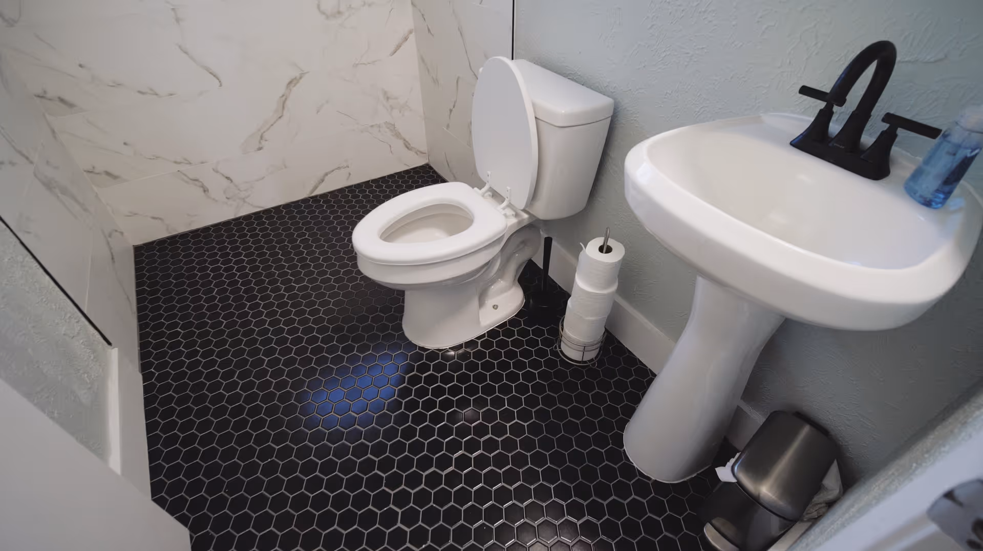 A small bathroom with a white toilet and a white pedestal sink. The floor is covered with black hexagonal tiles, and the walls have a light gray color with a section of white marble-patterned tiles. There is a toilet paper holder with multiple rolls next to the toilet, a soap dispenser on the sink, and a small trash can on the floor.