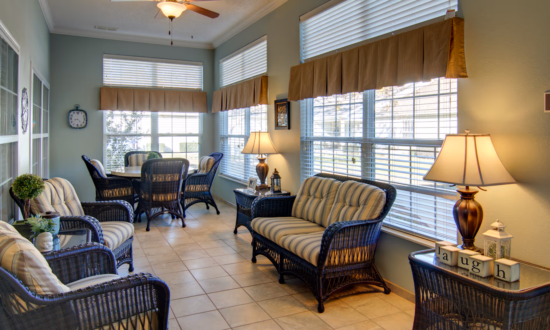 A bright sunroom with large windows covered by blinds and valances. The room is furnished with wicker chairs and a loveseat with striped cushions, a round table with four wicker chairs, two side tables with lamps, and decorative items including a small plant and blocks spelling 'laugh'. The floor is tiled and the walls are painted light blue.