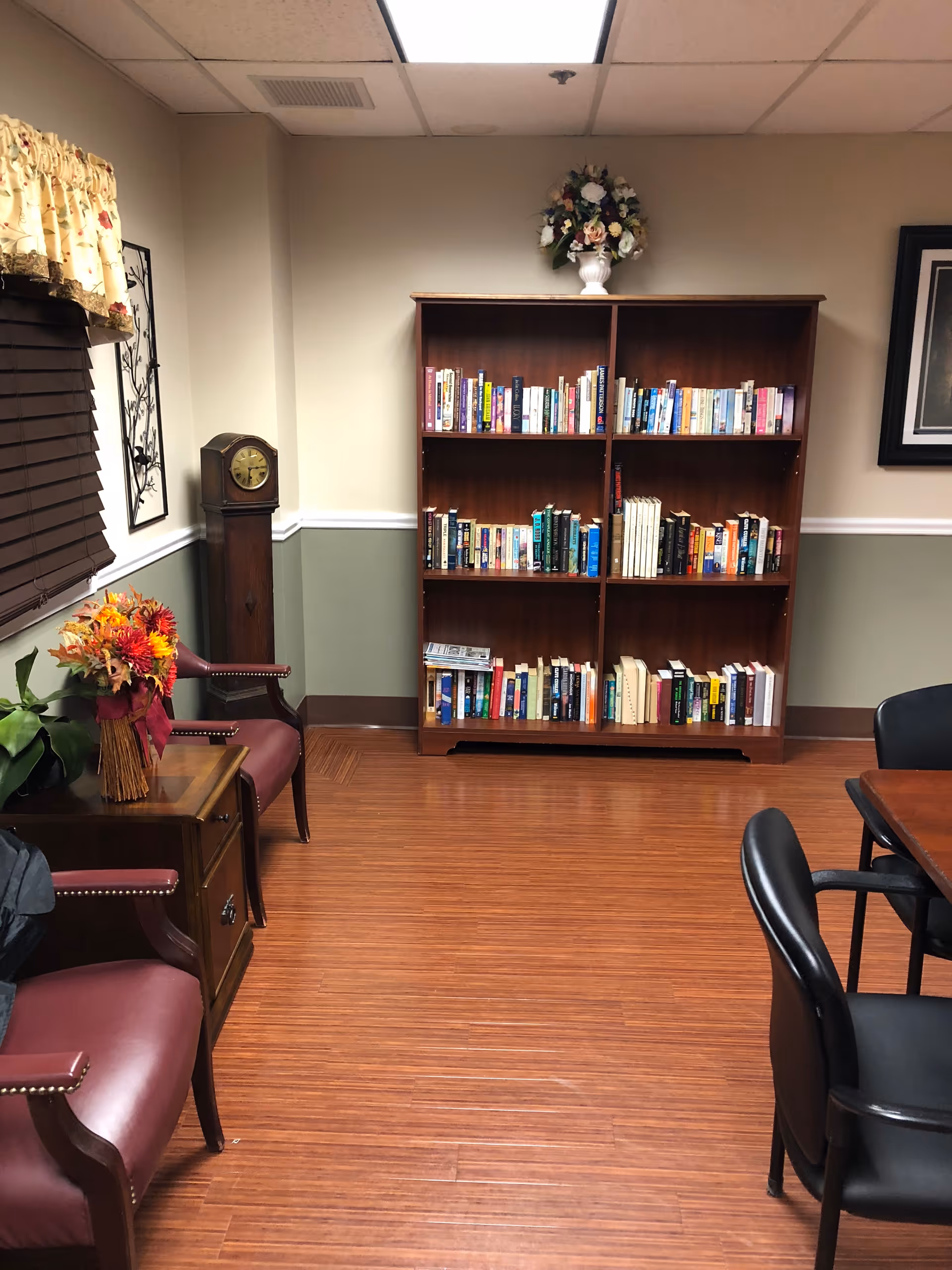 A cozy room with wooden flooring featuring a tall wooden bookshelf filled with books, a grandfather clock in the corner, two maroon leather chairs with nailhead trim, a small wooden side table with a floral arrangement, and a window with brown blinds and a floral valance. There is also a wooden table with black chairs partially visible on the right side.