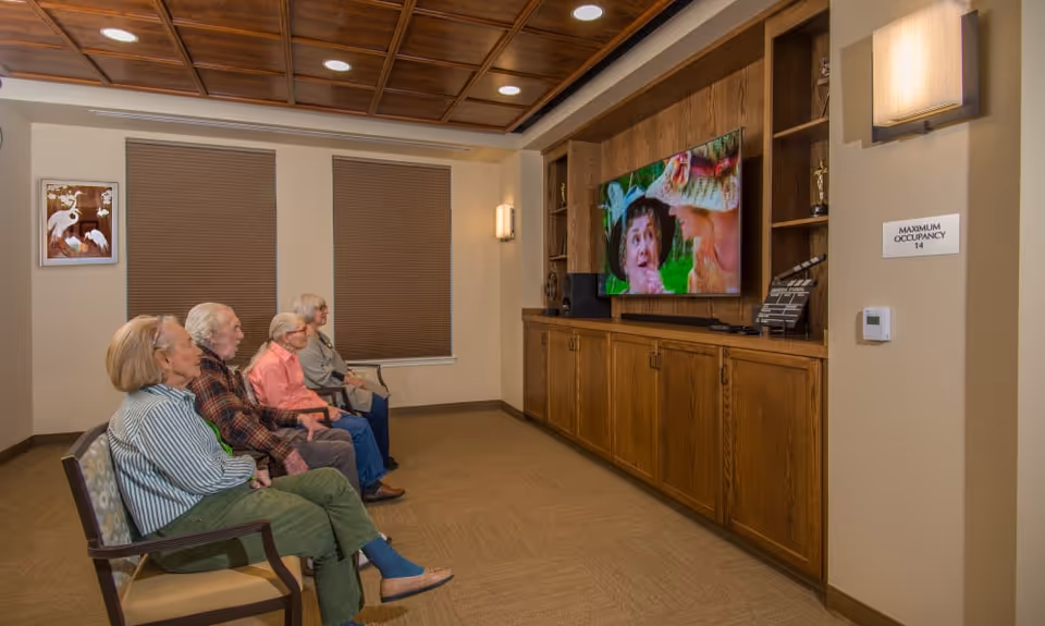 Four elderly people sitting in chairs in a room watching a movie on a large flat-screen TV mounted on a wooden cabinet. The room has beige walls, two windows with closed blinds, a wooden coffered ceiling, and a sign on the wall indicating a maximum occupancy of 14.