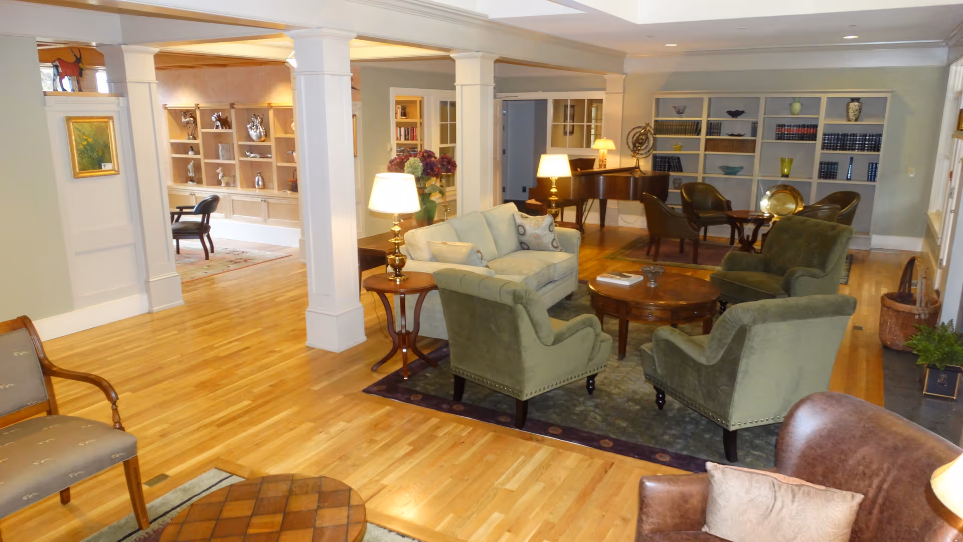 A spacious and well-lit living room area with wooden floors, green upholstered armchairs, a light-colored sofa, a round wooden coffee table, side tables with lamps, and built-in bookshelves filled with books and decorative items. There is also a piano in the background and additional seating areas visible through an open doorway.