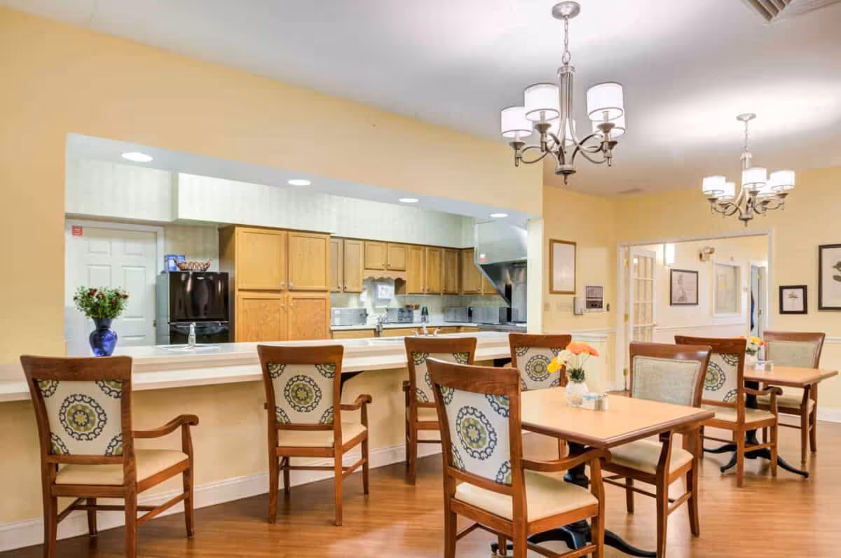 A dining area in a senior living facility featuring wooden tables and chairs with patterned upholstery. The room has warm yellow walls and hardwood floors. There is a counter with additional chairs facing an open kitchen area with wooden cabinets and stainless steel appliances. Two chandeliers hang from the ceiling providing light.