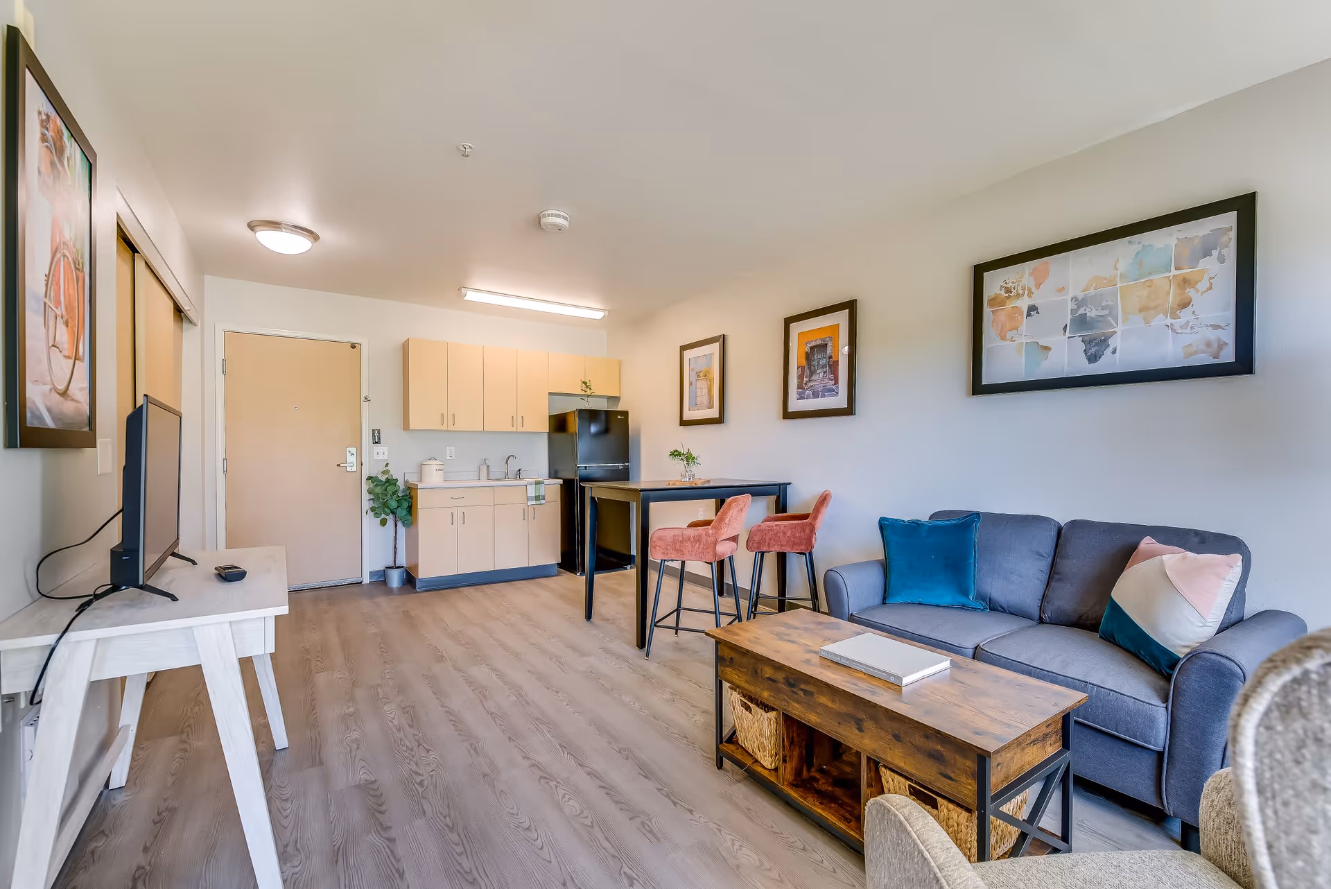 A bright and modern senior living apartment interior featuring a small kitchenette with light wood cabinets, a black refrigerator, and a sink. Adjacent to the kitchenette is a high table with two pink upholstered chairs. The living area includes a gray sofa with colorful pillows, a wooden coffee table with storage baskets underneath, and a TV on a white stand. The walls are decorated with framed artwork, including a world map and other pictures. The floor is light wood, and the entrance door is visible in the background.